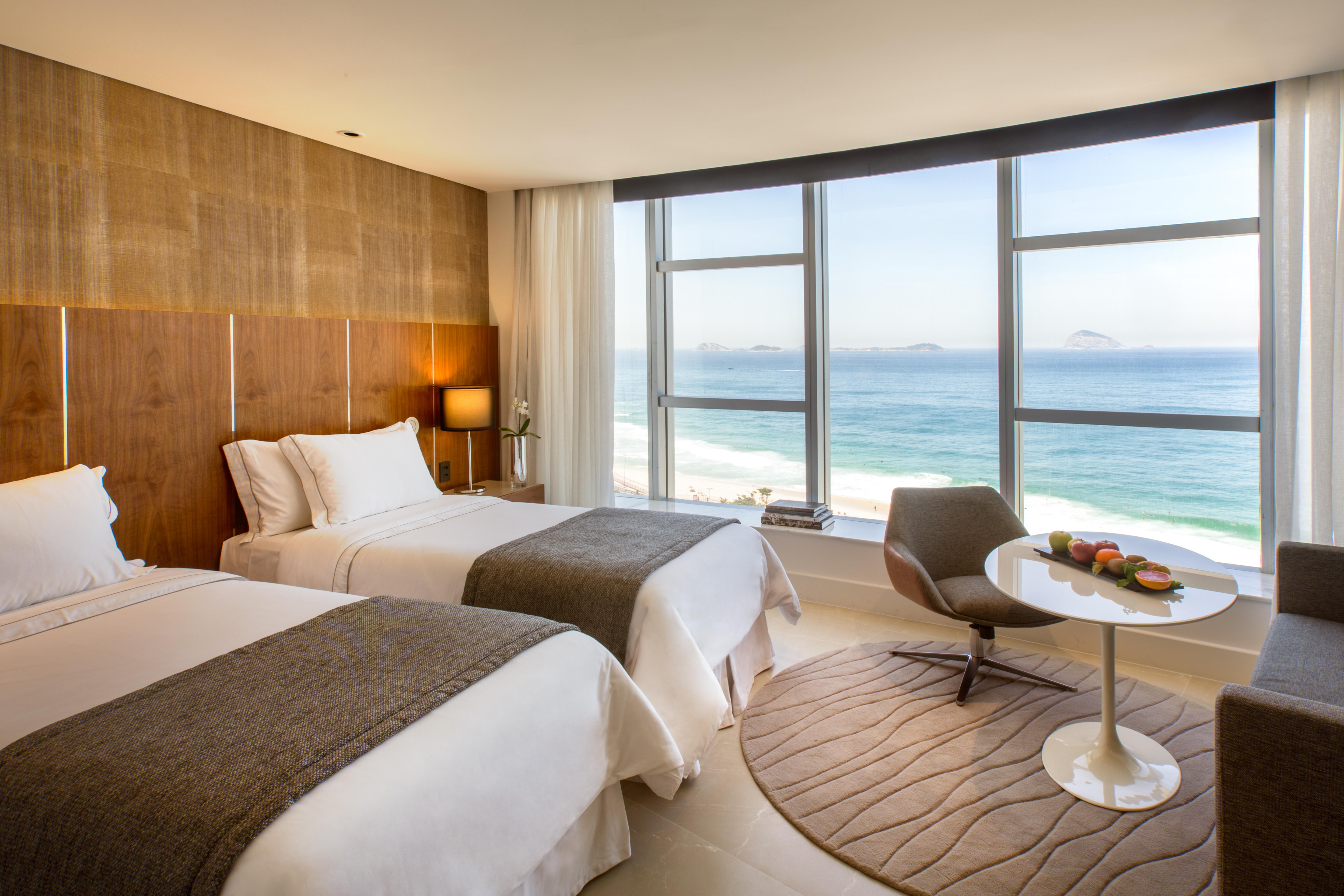 Hotel Nacional Rio De Janeiro - Oficial Ngoại thất bức ảnh The photo shows a modern hotel room with two beds adorned in white linens and gray throws. Large windows allow natural light to flood the space, offering a stunning view of the ocean. In the corner, there's a comfortable chair and a small round table